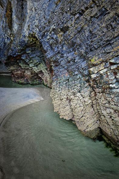 The shot captures a serene, sandy cove on the Donegal coast, leading into a calm, shallow pool of green-tinged water. On the right, a dramatically textured cliff face, composed of slanted layers of dark grey and blue rock, curves around the water. The left side of the image features an opposing rock formation, also layered, creating a natural archway. The sand shows ripples from the receding tide, and the overall mood is tranquil and contemplative, highlighting the rugged natural beauty of the Donegal coastline in Ireland.