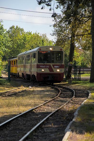 Pociąg osobowy o numerze MBxd2-212 wjeżdża na torowisko otoczone drzewami i zielenią. Na wyświetlaczu czołowym widnieje kierunek jazdy: 'Prawy Brzeg Wisły'. Za głównym wagonem ciągną się żółto-czerwone wagony doczepne. Scena rozgrywa się w słoneczny dzień w otoczeniu przyrody.

A passenger train numbered MBxd2-212 is entering a track surrounded by trees and greenery. The front display shows the destination: 'Prawy Brzeg Wisły' (Right Bank of the Vistula). Yellow and red carriages follow the main unit. The scene takes place on a sunny day amid a natural landscape.