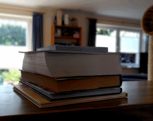 Photo von einem Bücherstapel, der meine lebende Bibliothek darstellen soll. Im Hintergrund weichgezeichnet ein Wohnzimmer mit einem Bücherregal