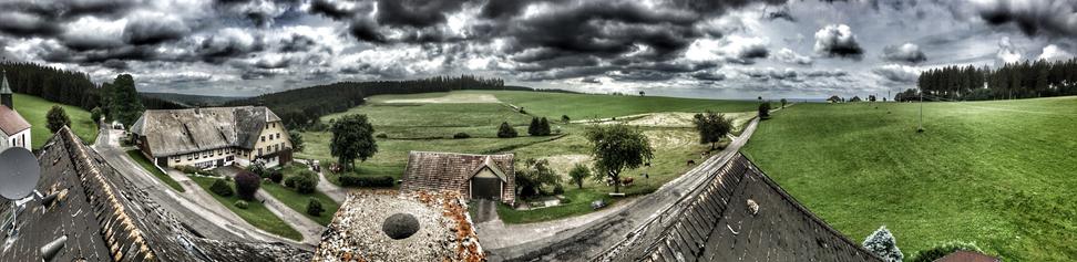 A panoramic view of a rural landscape featuring green fields, scattered trees, and a cloudy sky. Several buildings, including a large farmhouse, are visible along a winding road, with distant hills and forests in the background.