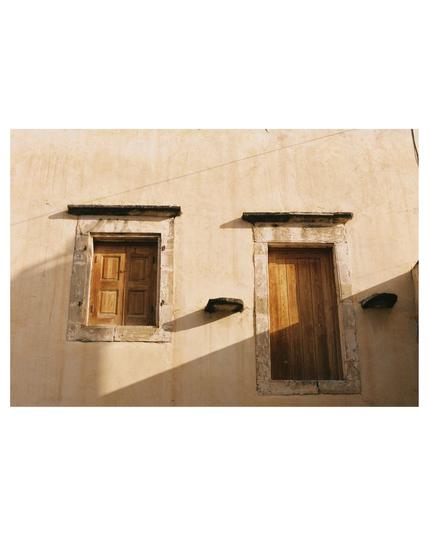 front of a building with a wooden closed window and a door. There is no stairway to the door, it starts midair. Morning light is coming from the right, long shadows shape the photo. Warm beige colors