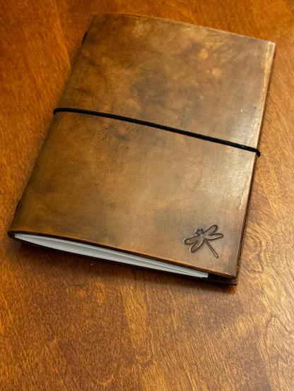 Handmade leather journal, with a glossy brown finish. It has a dragonfly stamped in the lower right corner, and is held shut with a horizontal black elastic