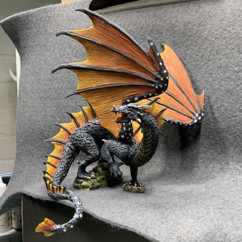 A black dragon with orange and yellow wings like a monarch butterfly. The wings are held upwards, and its tail wraps around its front. It has an open mouth with gore dripping from it.