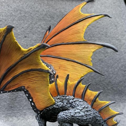 A black dragon with orange and yellow wings like a monarch butterfly. The wings are held upwards, and its tail wraps around its front. It has an open mouth with gore dripping from it.