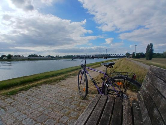 Blick vom Deich mit Wegen und Gras auf einen breiteren Fluß. Im Hintergrund eine Metallbrücke, vorne im Bild eine Pausenbank neben der ein Fahrrad steht. Blau-bewölkter Himmel.