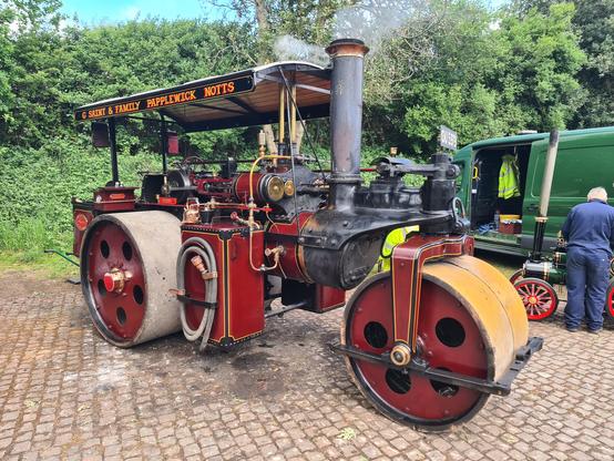 "G Saint & Family Papplewick Notts" traction engine.
The engine is idling, with steam coming out of the chimney.
The engine itself is painted a dark red, with a black roof.