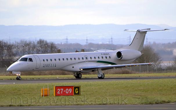 Side view of a twin engined jet airliner taxiing from right to left on a black runway.
The plane is almost entirely white, with a grey and green stripe swooping over the wing root.
There are green "Amelia" titles on the lower forward fuselage, and the black registration "F-HOXY" on the upper rear.
The foreground is filled with green grass, with a red sign with white numbers "27-09"  in the middle and the yellow letter "G" in a black square outlined in yellow in the centre of that grass.
Trees fill the background, with hills rising into hazy grey skies in the distance.