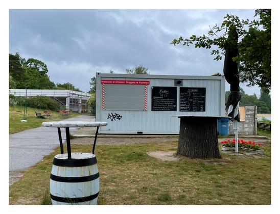 Photo taken in an outdoor pool on a rainy summer day: The snack bar in the center of the image is closed, empty bar tables looking like barrels. Two flower boxes of geraniums are standing under a folded parasol. Construction site in the background.