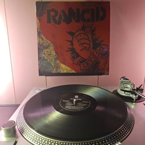 A vinyl record sits on a turntable. Behind the turntable, a vinyl album outer sleeve is displayed. The front cover shows a tattoo of a fist with "Let's Go" in a banner underneath, and a map of California.