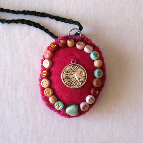 One side of the bright pink felt amulet with letter beads that say "neuro queer punk witch" and a charm of a sun in the middle.