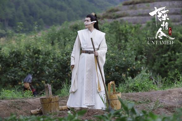 A wider shot of a farming area, centered on a standing Jin Zixuan who is looking beyond the left side of the image. A bucket sits on the ground to either sets if him, a carry pole lying on the ground by one bucket. A thin cloth is tied around his head to cover his nostrils, and he holds a long pole in his left hand. Tucked into the left side of the image is the out of focus head and shoulders of a Wen guard.