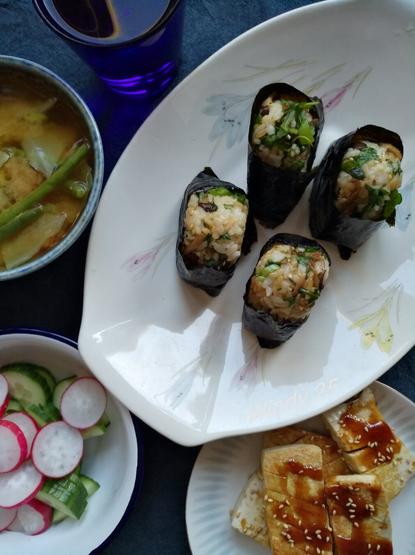 Four dishes, one with the riceballs, one small miso soup, one with slices of cucumber and radish, one with tofu. Also cobolt blue drinking glass partly in the picture.