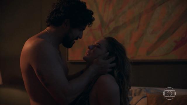 A imagem mostra uma cena de um casal em um momento íntimo e emotivo. O homem está sem camisa, segurando delicadamente o rosto da mulher com as duas mãos, enquanto ambos se encaram intensamente. A mulher, com expressão emocionada, segura nos braços dele. Ao fundo, há uma pintura abstrata com tons de laranja, verde e vermelho, e parte de uma cama com almofadas visível. No canto inferior direito da imagem, aparece a logomarca da TV Globo, indicando que a cena é de uma produção da emissora. A iluminação é suave e quente, criando um clima íntimo e acolhedor.