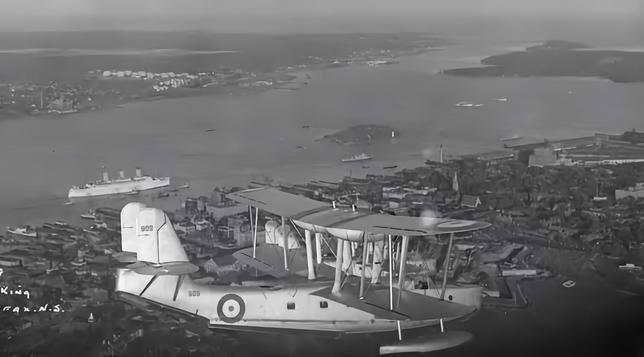 A biplane flying boat seen in a three-quarter vie from the straboard side. The background shows a city, abnd beyond it an ocean harbour with ships visible.