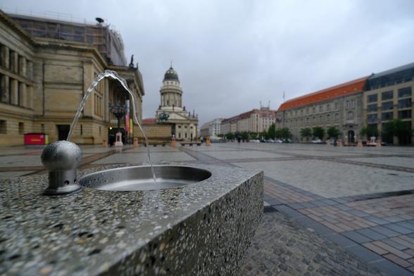 The image shows a city square (Gendarmenmarkt) with a drinking fountain in the foreground. The square is wet, likely from recent rain, and features historic buildings in the background, including one with a notable dome. The scene is calm and slightly overcast. The photo appears to be taken with a wide-angle lens, capturing a broad view of the square and its surroundings.