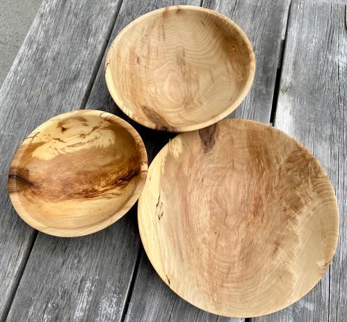 Three small bowls from a golden brown wood sitting on grey cedar planks outside.