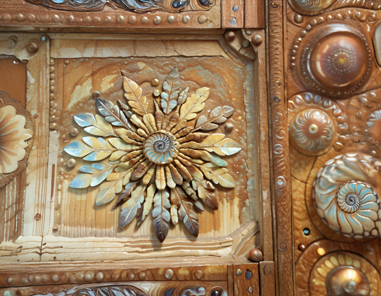 An intricately carved panel featuring a loose flower design of feather-like petals and a central spiral ammonite motif in shades of brown, gold, tan, and turquoise.