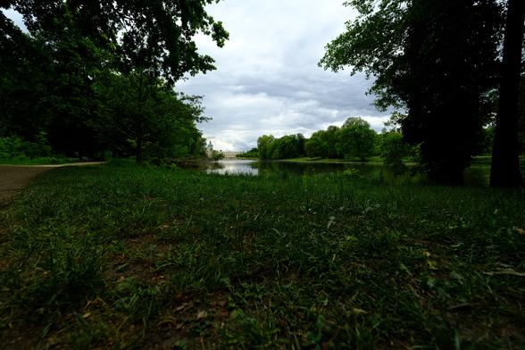 The image depicts a serene park scene with a path leading through lush greenery towards a body of water. In the distance, a grand building with a white facade and green roof is visible (Schloss Charlottenburg), framed by trees under a cloudy sky. The photo is taken from a low angle, emphasizing the grass and trees in the foreground.