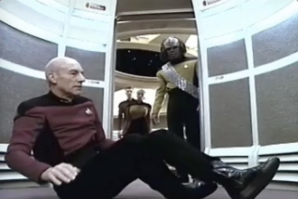 Picard sits on the floor of the turbolift. The turbolift doors are open to show Worf and two other members of the bridge crew checking on him to see if he is okay.