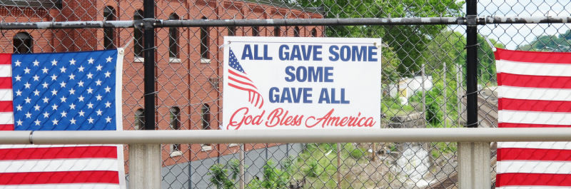 An einem Maschendrahtzaun mit Klinkerhaus im Hintergrund sind
zwei US-Fahnen angebracht, dazwischen ein Schild mit der Aufschrift:
„All gave some/Some gave all/God bless America“.