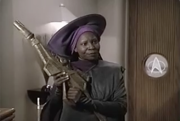 Guinan stands in Ten Forward holding a gold phaser in her hands after firing it inside to defuse a tense situation.