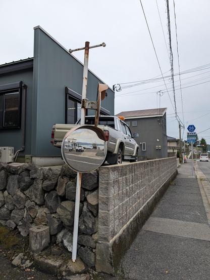 The seaside can be seen reflected on a road mirror opposite to the water. Behind the roadmirror there is a wall supporting an elevated house and a pickup truck.