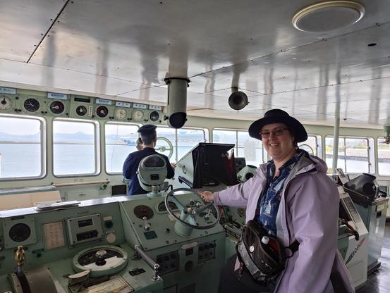 A photo of a caucasian woman in a lavender raincoat and broad hat on the helm of an antique ship.