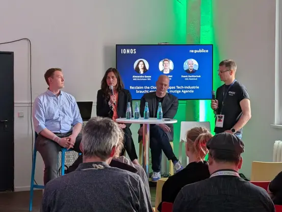 Arthur Mai, Alexandra Geese, Frank Karlitschek sitzen um einen Tischr vor einem Bildschirm mit blauem Hintergrund, Andreas Maurer steht mit einem Mikrofon in der Hand daneben.
