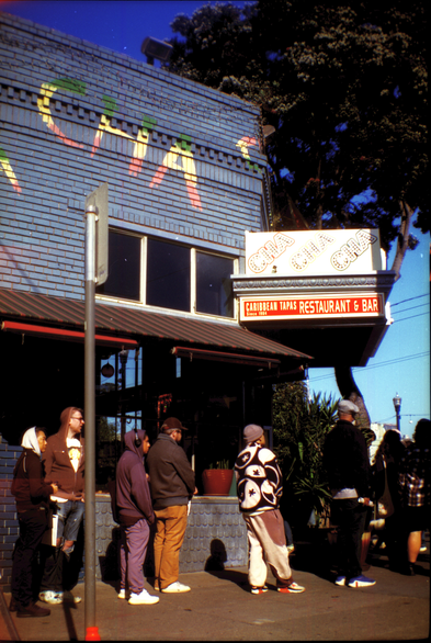 people lined up around the block to buy records, rounding a corner with a brick building painted kind of blue, with a rainbowy sign saying "Cha" (I think the last part of Cha Cha Cha)