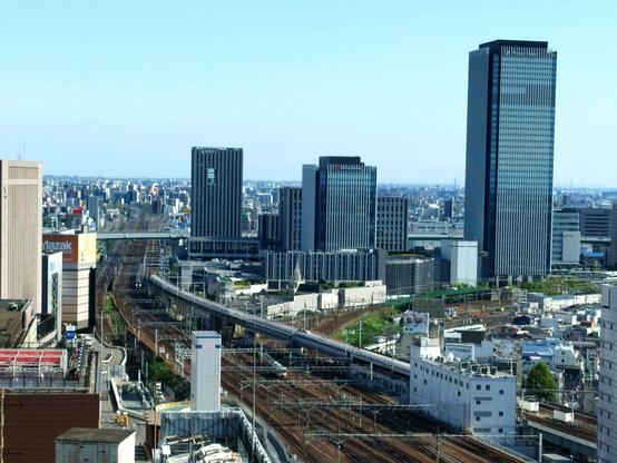 Picture taken from one of the skyscrapers sitting on top of Nagoya station looking out slightly from the side at the bundle of train tracks that cut through the city from here going southward. There is a single white bullet train just starting to speed up. Most of it is in the shot. There are two more conventional and much shorter trains also. And a but of the skyline left and right of it all.