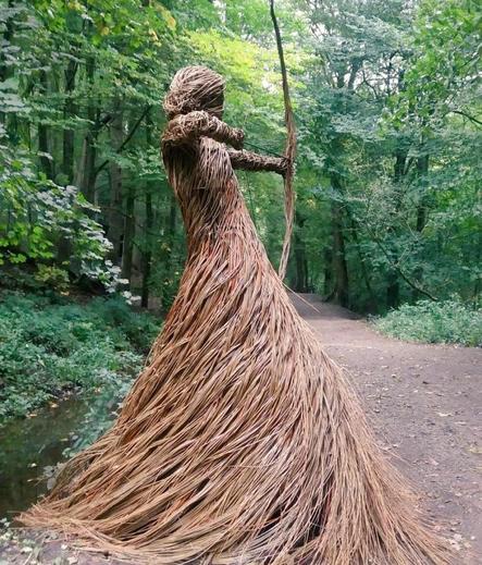 Photo of a brown willow sculpture of a female archer holding a bow as if about to release an arrow, with hair tied back and in a long flowing skirt, the sculpture is on a path in a green woodland setting