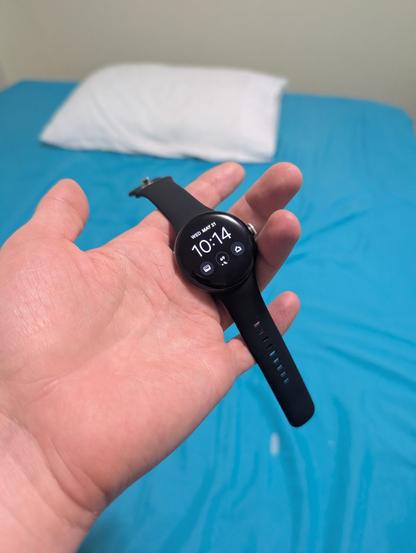 A Pixel Watch is held in a man's hand. The time, 10:14 appears on the face.