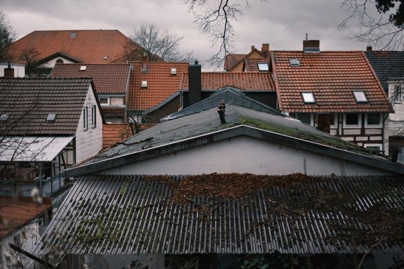 Blick auf Dächer in einem Wohngebiet mit verschiedenen architektonischen Stilen. Einige Dächer haben rote Ziegel, während andere flach und mit Moos und Blättern bedeckt sind. Der Himmel ist bedeckt, was zu einer stimmungsvollen Atmosphäre beiträgt.
