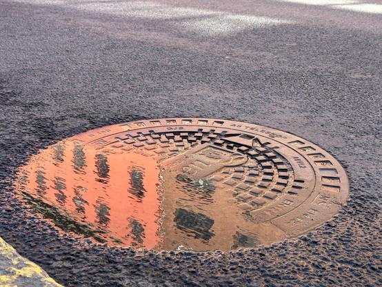 A circular utility hole cover on a road. It is slightly inclined, and about half of it is covered with water, which reflects two buildings (one red, one tan)