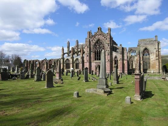 Melrose Abbey. The image shows a side view of the length of the abbey and  abbey church at Melrose, with the taller ruins of the church on the right and other elements on the left. Leading back to the abbey is a broad area of mown grass containing gravestones and monuments. The scene is in sunshine.