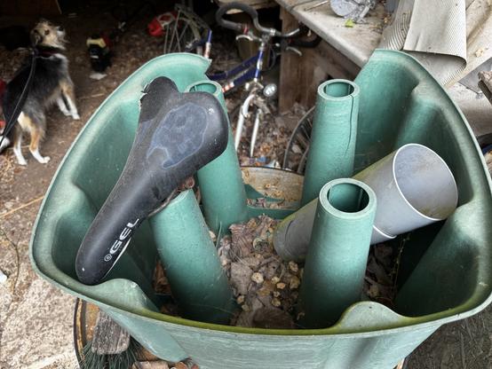 Eine unordentliche Szene in einem Schuppen mit einem grünen Plastikbehälter, der mit Blättern und undefiniertem Zeug gefüllt ist, verschiedenen grünen Rohren, die aus ihm hervorstehen, und einem Fahrradsitz, der darauf ruht. Im Hintergrund sind Fahrradreste und ein Hund zu sehen, der neugierig auf die Szene schaut.