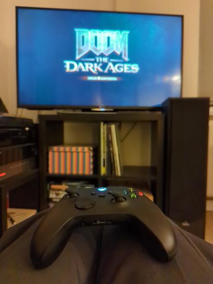Im Vordergrund sieht man einen XBox-Controller, um Hintergrund zeigt ein Fernseher das Start Logo von "Doom: The Dark Ages"