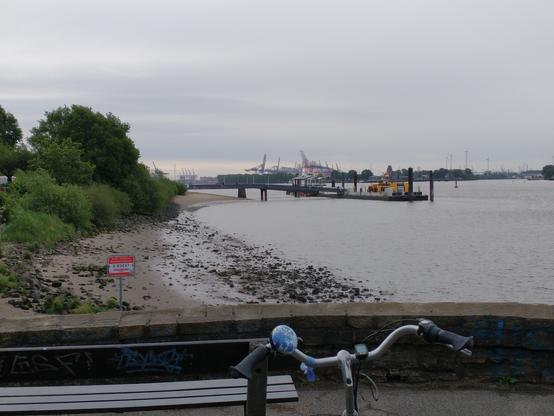Man sieht die Elbe und den Fähranleger Teufelsbrück. Himmel und Wasser sind grau. Am linken Rand ist ein Stück Ufer mit Sand und Steinen zu sehen, daneben Sträucher. Im Vordergrund ragen ein Fahrradlenker und eine kleine Mauer ins Bild.
