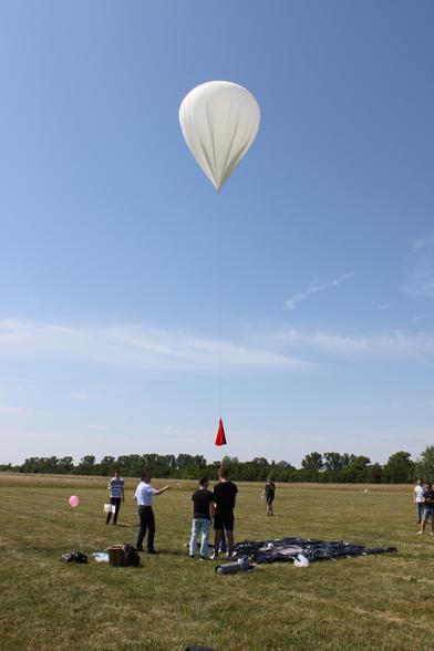 A group of people launching a white weather balloon on a large lawn, blue sky and some trees in the far back