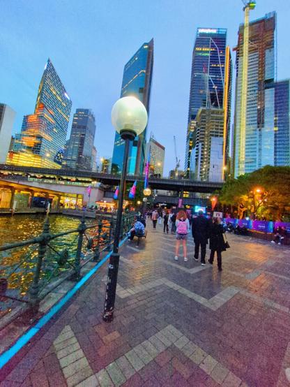 People walking along the waterfront at Circular Quay. To the left, shimmering golden light reflects off the water of Sydney Harbour. Tall blue glass skyscrapers stand on the horizon.