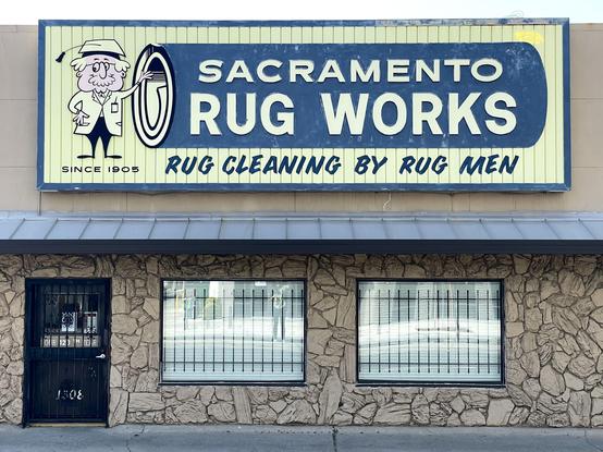 Photo of a sign on a building. A cartoon man with a mortarboard cap stands next to a rug with the words: “Sacramento Rug Works”. Also on the sign:

SINCE 1905
RUG CLEANING BY RUG MEN