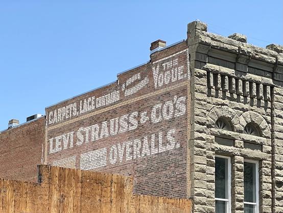 Photo of a ghost sign on the side of a stone building. It advertises “Carpets, Lace Curtains, The Vogue, Levi Strauss & Co’s Overalls.”