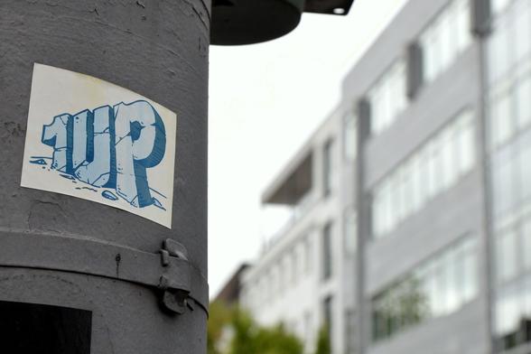 1UP auf einem Aufkleber, der an einem dunkelgrüngrauen Laternenpfahl klebt auf der linken Seite. Rechts und im Hintergrund ist unscharf ein graues mehrgeschossiges Bürogebäude zu sehen.