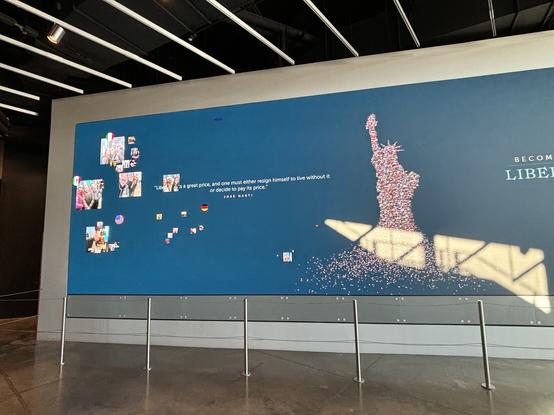 Großer Bildschirm, circa 3 m hoch und 6 m breit, mit blauem Hintergrund. Von links schweben Portraitbilder und Flaggen ein, die nach rechts kleiner werden und mit einer stilisiertes Miss Liberty verschmelzen.