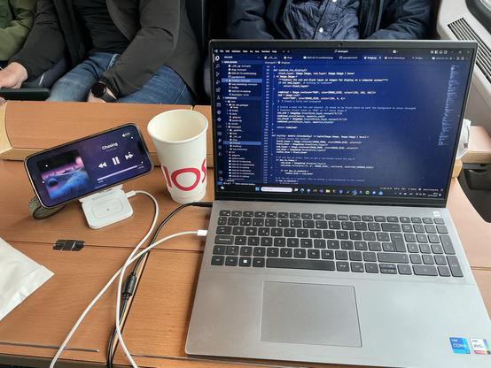 Table shot showing my laptop that is running a VSCode window. Next to it I have my paper cup of Kiosk tea, and my iPhone on a MagSafe charger in horizontal mode showing the current song: Chasing by Foudroie