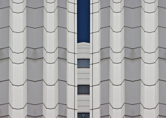 A photomontage of parts of a white building. Left and right of the photo vertical curtains (with folds) of stone. In the middle an opening through which you see three white horizontal stripes with blue windows in between and above that a strip of blue sky.