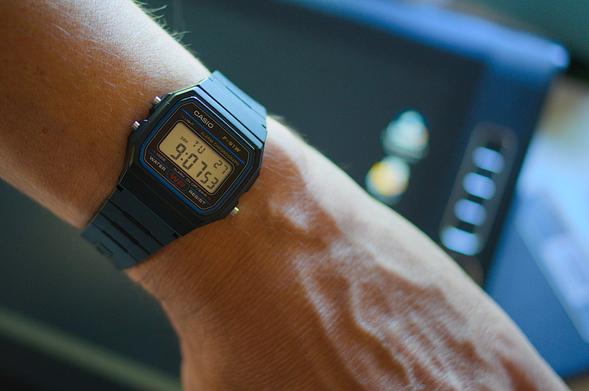 Fotografía donde se ve en primer plano un reloj Casio F-91W colocado en la muñeca de mi mano. De fondo un ordenador de pantalla táctil y algunas cintas de cassette.