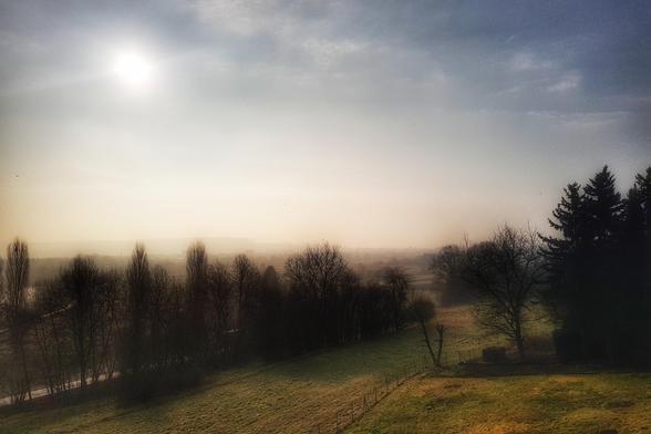 A wide shot of a misty, sun-drenched rural landscape in Germany during early morning. The sun, a bright circle, is visible in the upper left, casting a warm glow over the scene. A gently sloping grassy hill with a fence runs across the foreground and midground, leading to a dark line of bare trees. Beyond the trees, the mist obscures the distant view of what appears to be a town or village. The sky transitions from bright and hazy near the sun to a darker, more dramatic blue on the right.