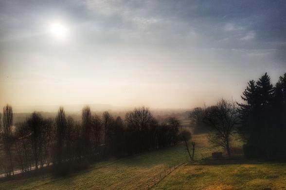 A wide shot of a misty, sun-drenched rural landscape in Germany during early morning. The sun, a bright circle, is visible in the upper left, casting a warm glow over the scene. A gently sloping grassy hill with a fence runs across the foreground and midground, leading to a dark line of bare trees. Beyond the trees, the mist obscures the distant view of what appears to be a town or village. The sky transitions from bright and hazy near the sun to a darker, more dramatic blue on the right.
