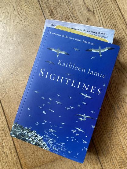 A book titled "Sightlines" by Kathleen Jamie is displayed against a wooden surface. The cover features a blue background with several white birds in flight. A piece of paper with text is partially visible behind the book.